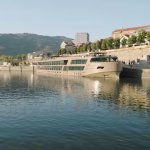 Beige river cruise ship moored at a waterfront promenade with trees and hills in the background.