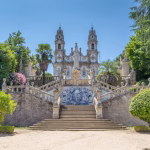Grand stone staircase leads up to a baroque church with twin towers, framed by lush trees; a blue tiled alcove sits at the landing.
