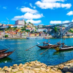 Porto waterfront with colorful hillside houses, Douro River, and the arched Dom Luís I Bridge. Boats float near the quay.