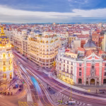 Bird's-eye view of a European city with ornate historic buildings along a curved street and light-trail traffic under a blue sky.