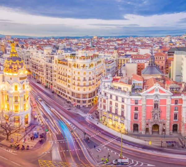 Bird's-eye view of a European city with ornate historic buildings along a curved street and light-trail traffic under a blue sky.