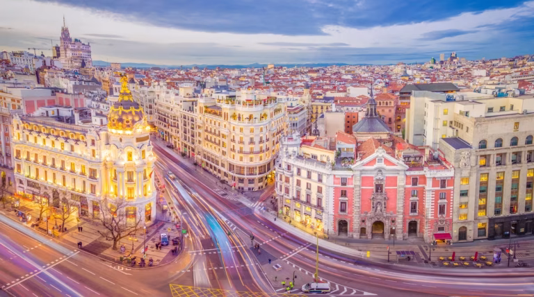 Bird's-eye view of a European city with ornate historic buildings along a curved street and light-trail traffic under a blue sky.