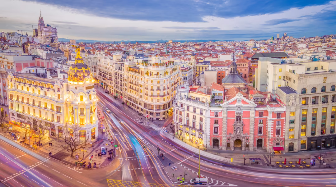 Bird's-eye view of a European city with ornate historic buildings along a curved street and light-trail traffic under a blue sky.