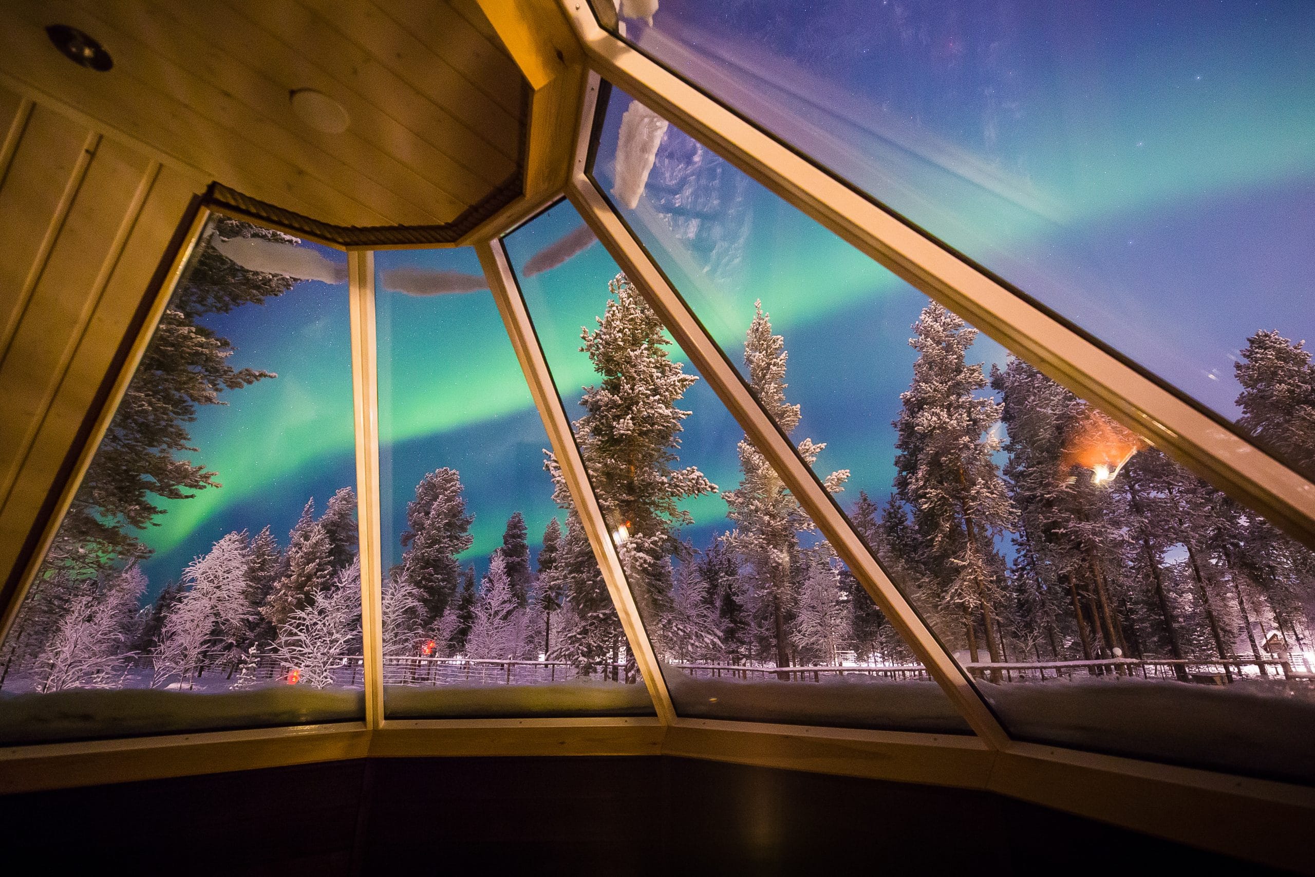 View through a glass-walled pavilion at night: snow-covered trees outside and the green aurora borealis in the sky behind the glass panes.