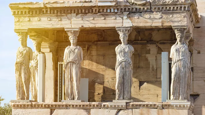 Four carved stone Caryatid columns supporting a temple balcony, bathed in warm sunlight on an ancient Greek structure.