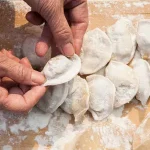 Hands shaping crescent dumplings on a floured wooden surface, with rows of uncooked dumplings ready to cook.