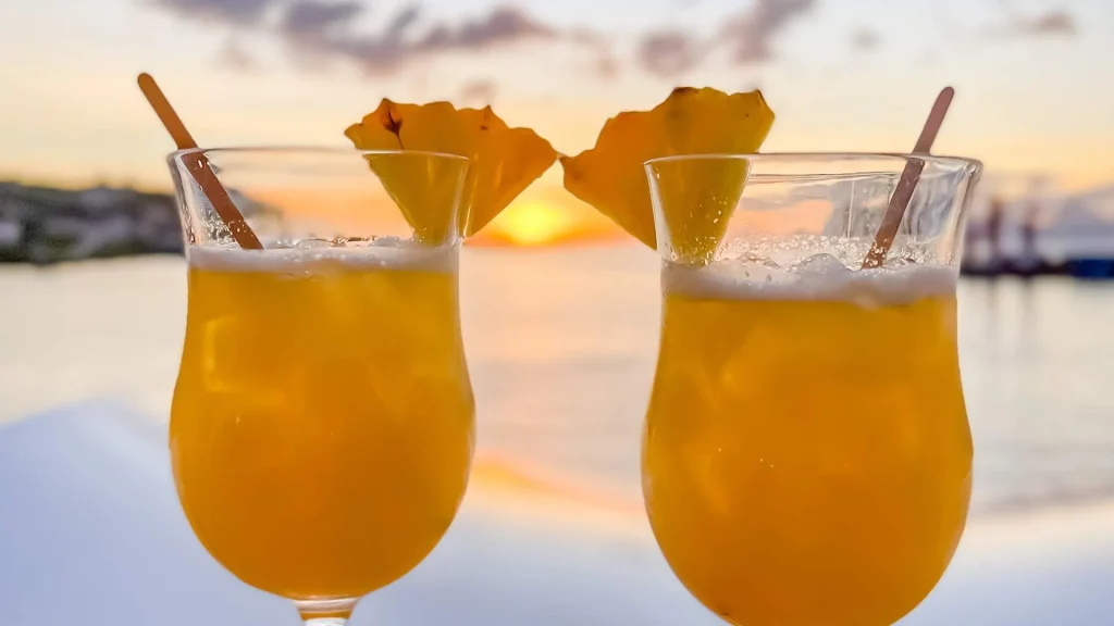 Two tropical orange cocktails in stemmed glasses with pineapple wedges and straws, set against a sunset backdrop.