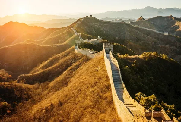 Section of the Great Wall winding over sunlit hills at sunrise, with distant watchtowers along the ridge.