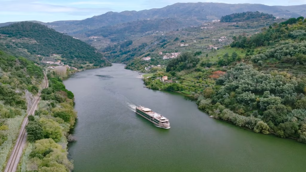 Passenger boat glides down a wide river through a lush green valley bordered by forested hills and small hillside houses amid the scenery.