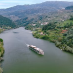 Passenger boat glides down a wide river through a lush green valley bordered by forested hills and small hillside houses amid the scenery.