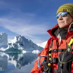 Person in a red life jacket and green knit cap stands beside icy water with icebergs and a blue sky in the background.
