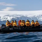 A group of people in yellow jackets sits on a black inflatable boat wearing life vests, navigating icy Antarctic waters with large icebergs in the background.