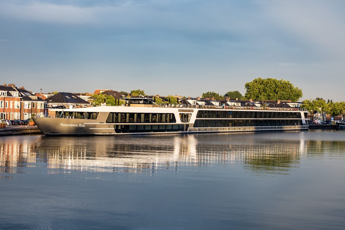 Modern river cruise ship TravelMarvel Rigel moored along a city waterfront with brick buildings and trees under a clear blue sky.