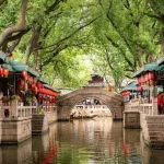 Canal market lined with stalls and red lanterns, a stone arched bridge in the distance over calm water under leafy trees.