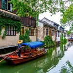 Man in a straw hat steers a wooden boat with a blue canopy along a canal lined with ivy-covered traditional buildings.
