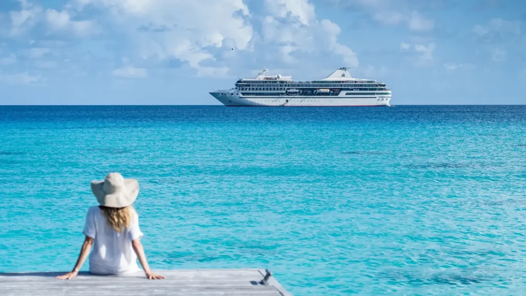 Woman in a sunhat sitting on a dock overlooking turquoise water, with a white cruise ship offshore.