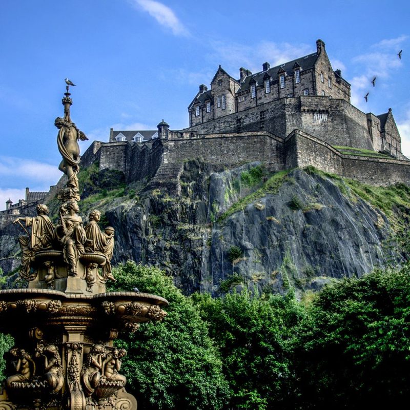 Castle atop rocky hill with a fortified stone wall, overlooking a lush green park.