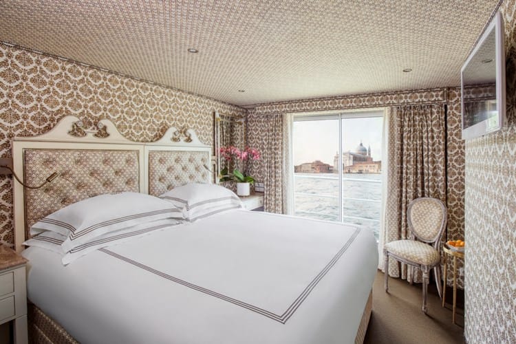 Luxurious hotel bedroom with tufted headboard, white bedding, ornate wallpaper, and a window with a water view.