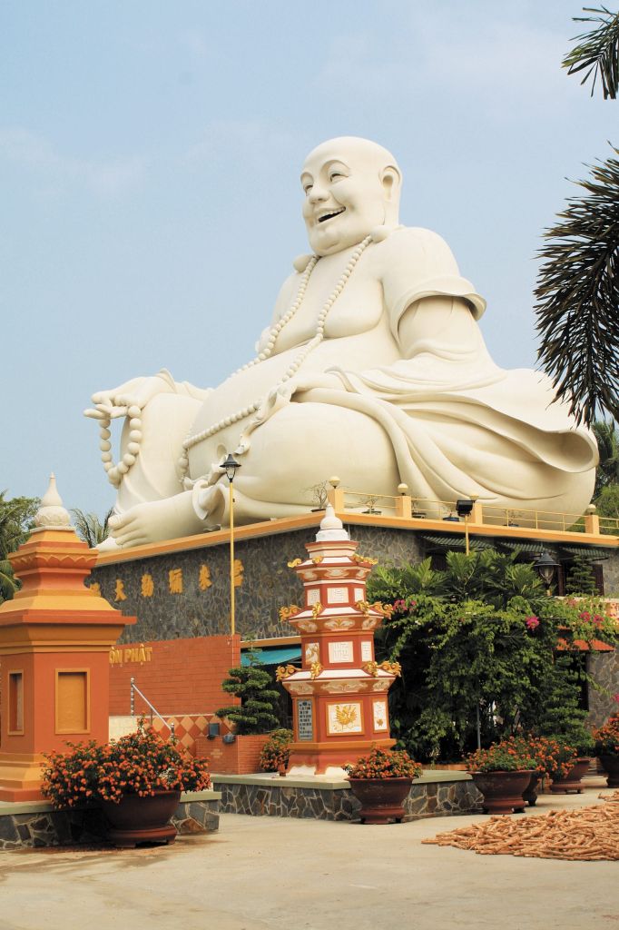 Large white statue of a Laughing Buddha seated in a temple courtyard with prayer beads