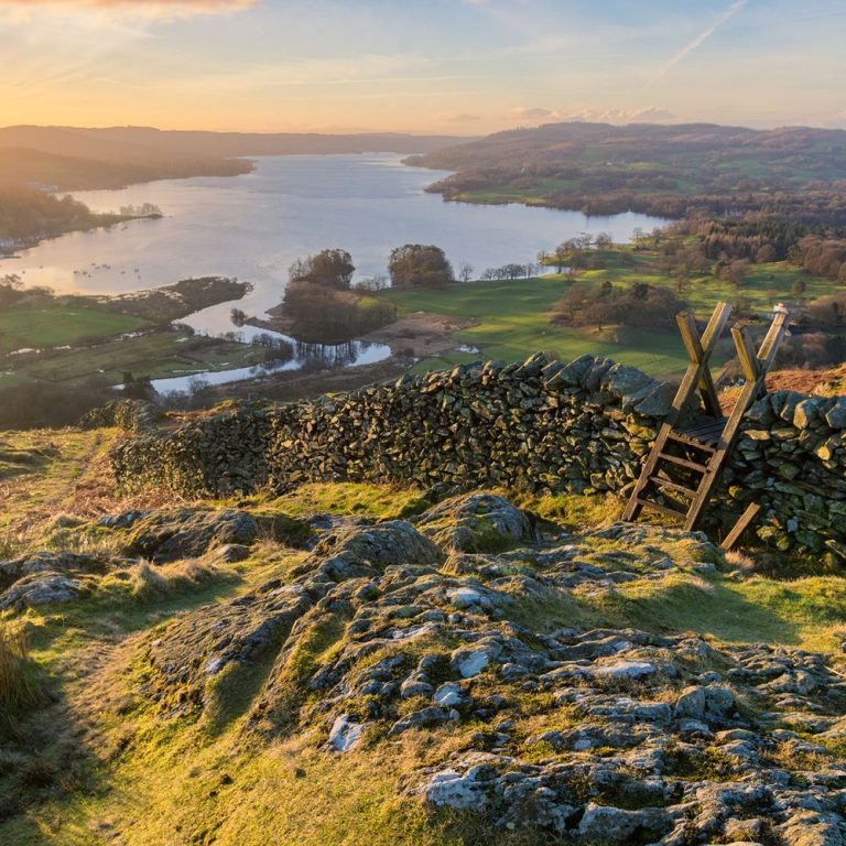 Sunlit countryside view from a rocky hill: a dry stone wall with a wooden stile leading to green fields and a winding river valley at sunset.