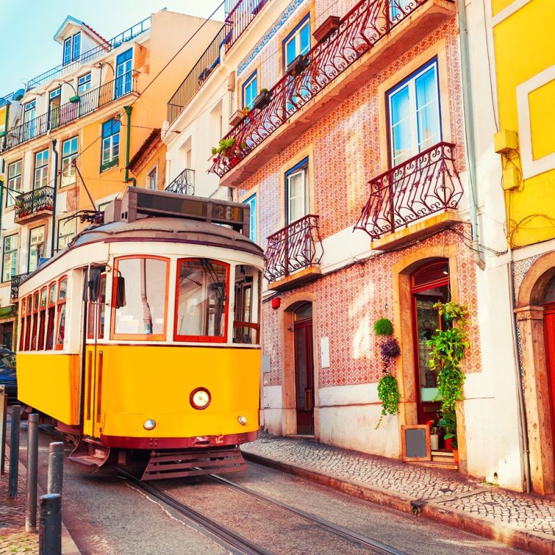 Yellow tram on a narrow cobblestone street beside colorful, tiled buildings with wrought-iron balconies.