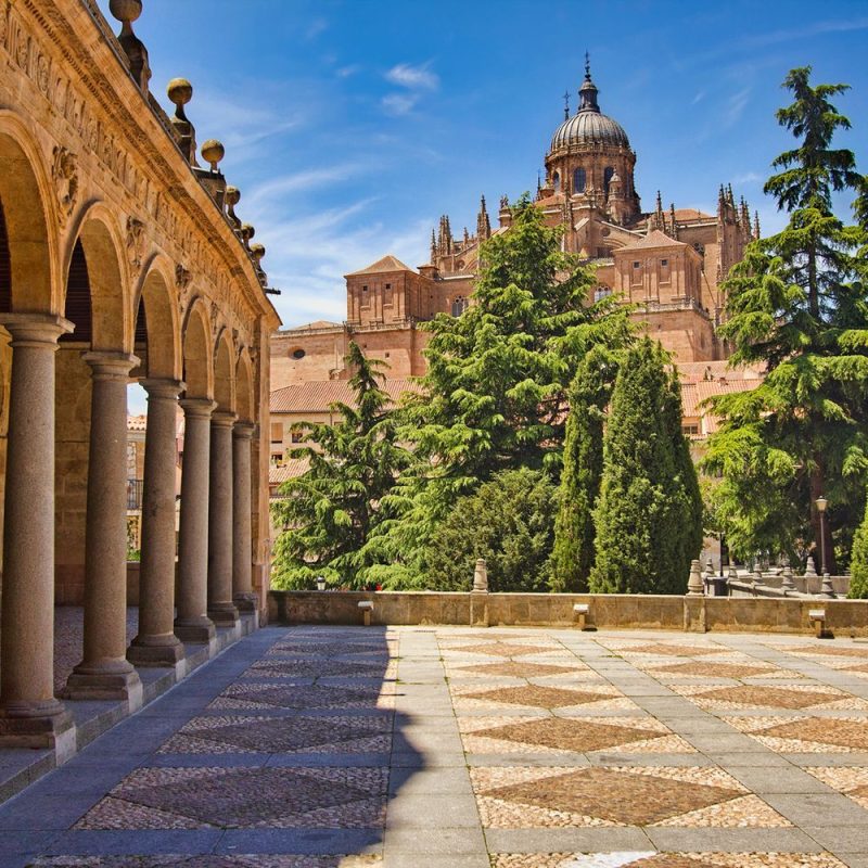 Stone colonnade on the left with arches, a tiled courtyard, and a domed cathedral rising behind green trees on a sunny day