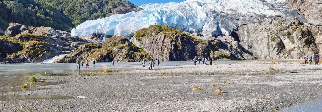 Hikers on a gravel shore in front of a blue glacier tongue in a rocky fjord landscape.