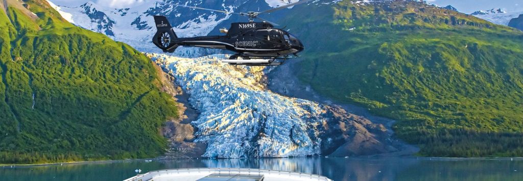 Black helicopter hovering over a glacier with a ship's deck in the foreground.