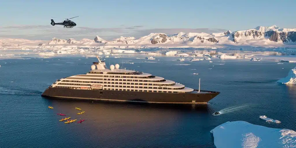 Expedition cruise ship with kayakers and a helicopter over icy Antarctic waters and snowy peaks.