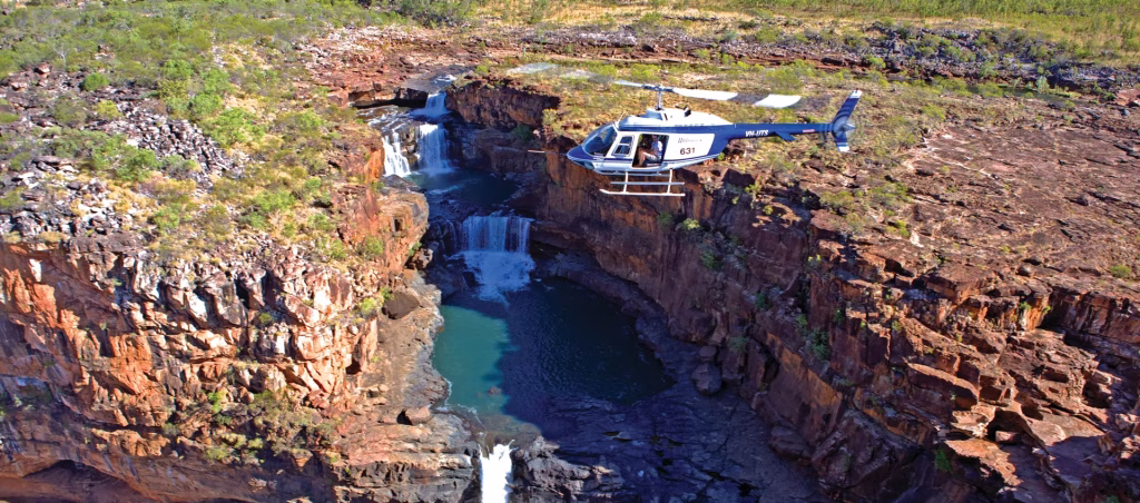 Helicopter flying over a red rock canyon with a waterfall and turquoise pool below.