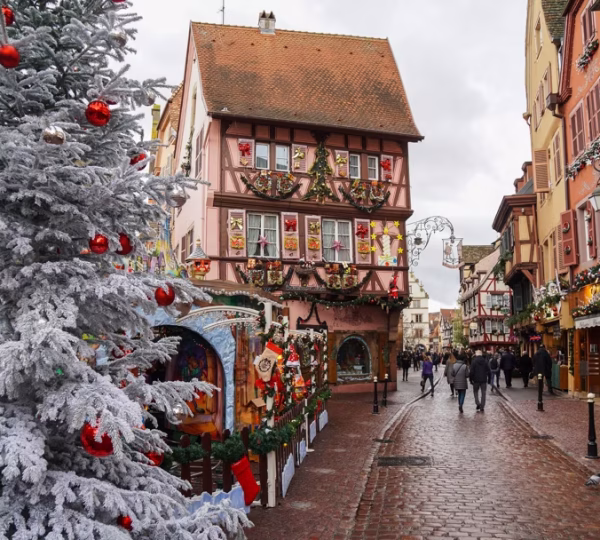 Snowy Christmas street with a frosted pine tree and colorful, ornamented houses along a cobblestone road, shoppers in the distance.
