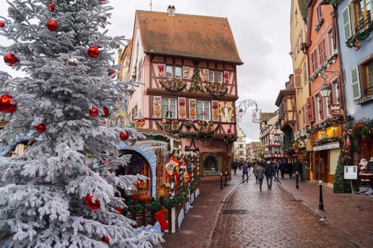 Snowy Christmas street with a frosted pine tree and colorful, ornamented houses along a cobblestone road, shoppers in the distance.