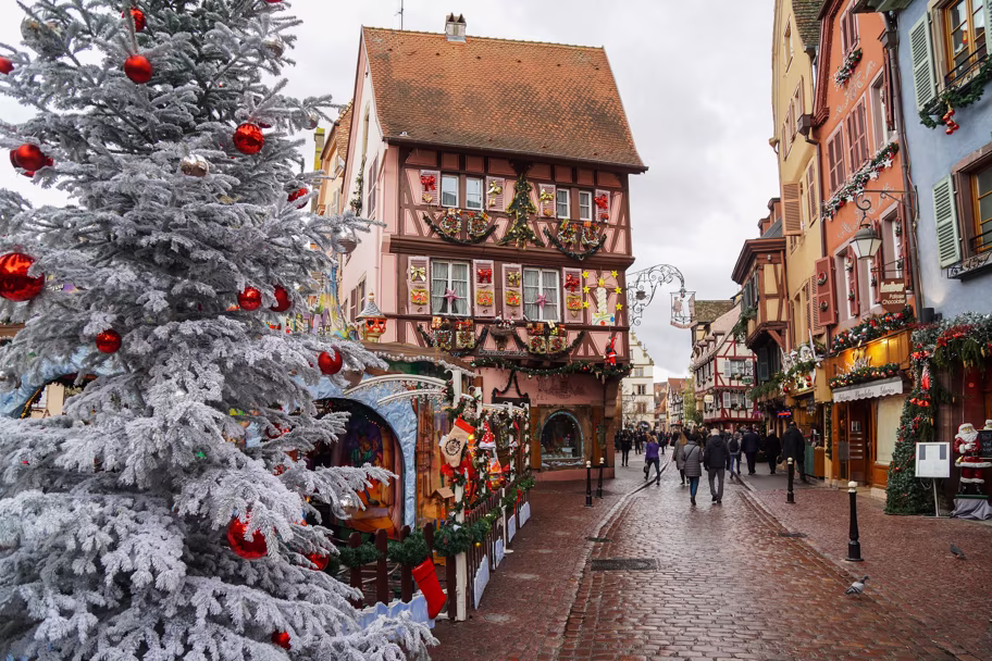 Snowy Christmas street with a frosted pine tree and colorful, ornamented houses along a cobblestone road, shoppers in the distance.