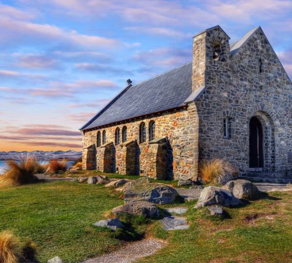 Stone church with a small bell tower and arched doorways on a grassy lakeside bluff, mountains in the distance at sunset.