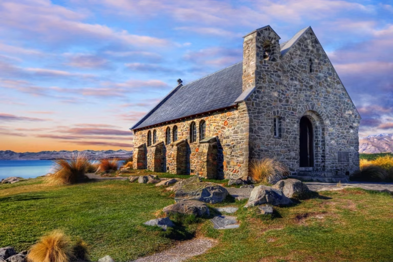Stone church with a small bell tower and arched doorways on a grassy lakeside bluff, mountains in the distance at sunset.