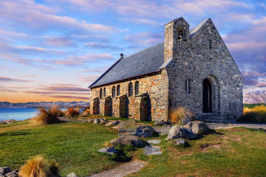 Stone church with a small bell tower and arched doorways on a grassy lakeside bluff, mountains in the distance at sunset.