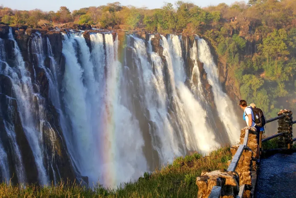 Two visitors at a stone railing overlooking a large waterfall with a rainbow in the spray.