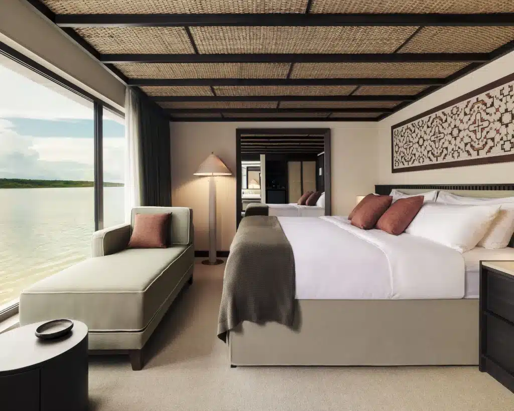Ship suite with king bed, chaise longue and window view across calm water.