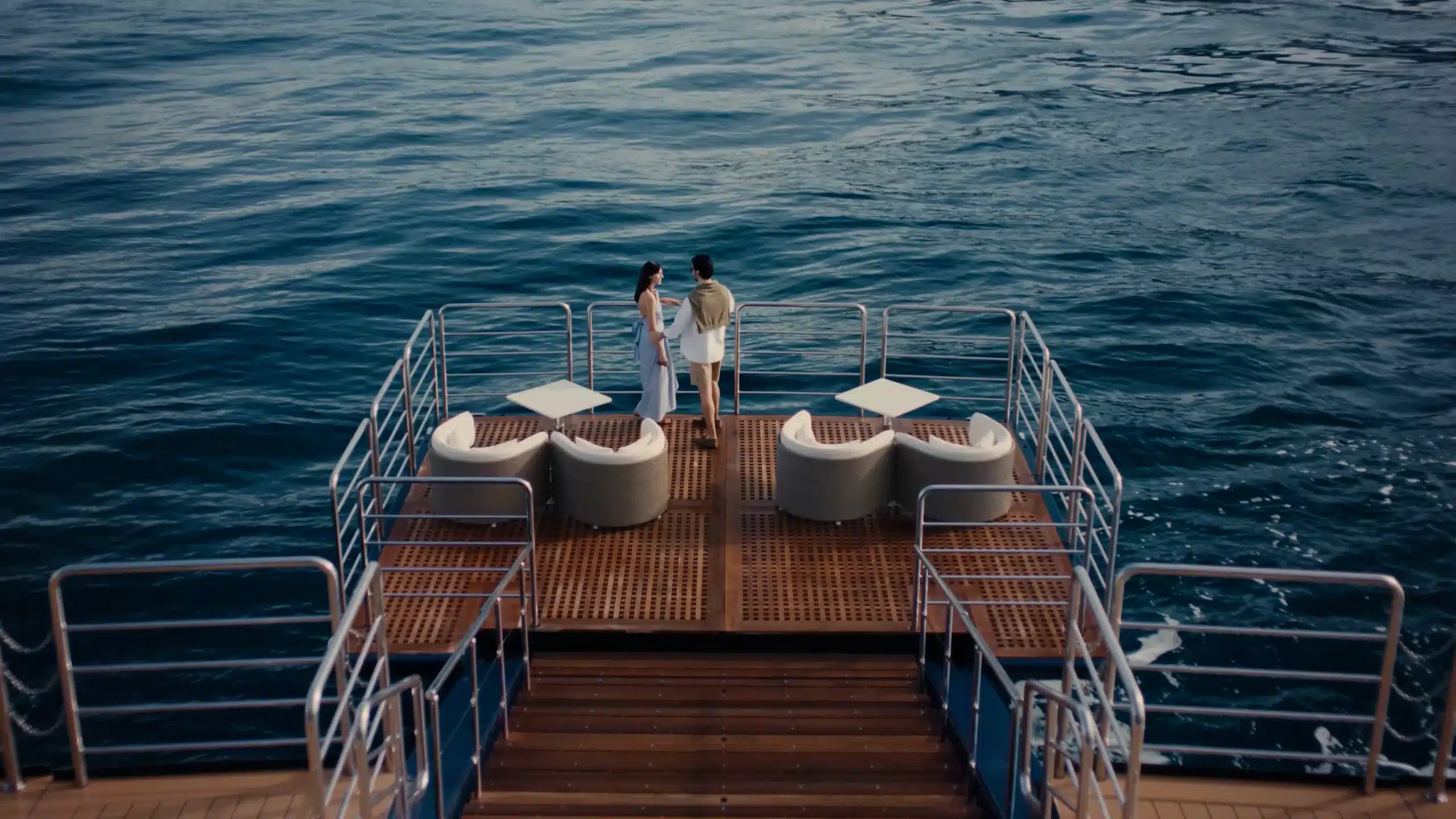Couple standing on a teak deck at the stern of a yacht overlooking open blue sea.