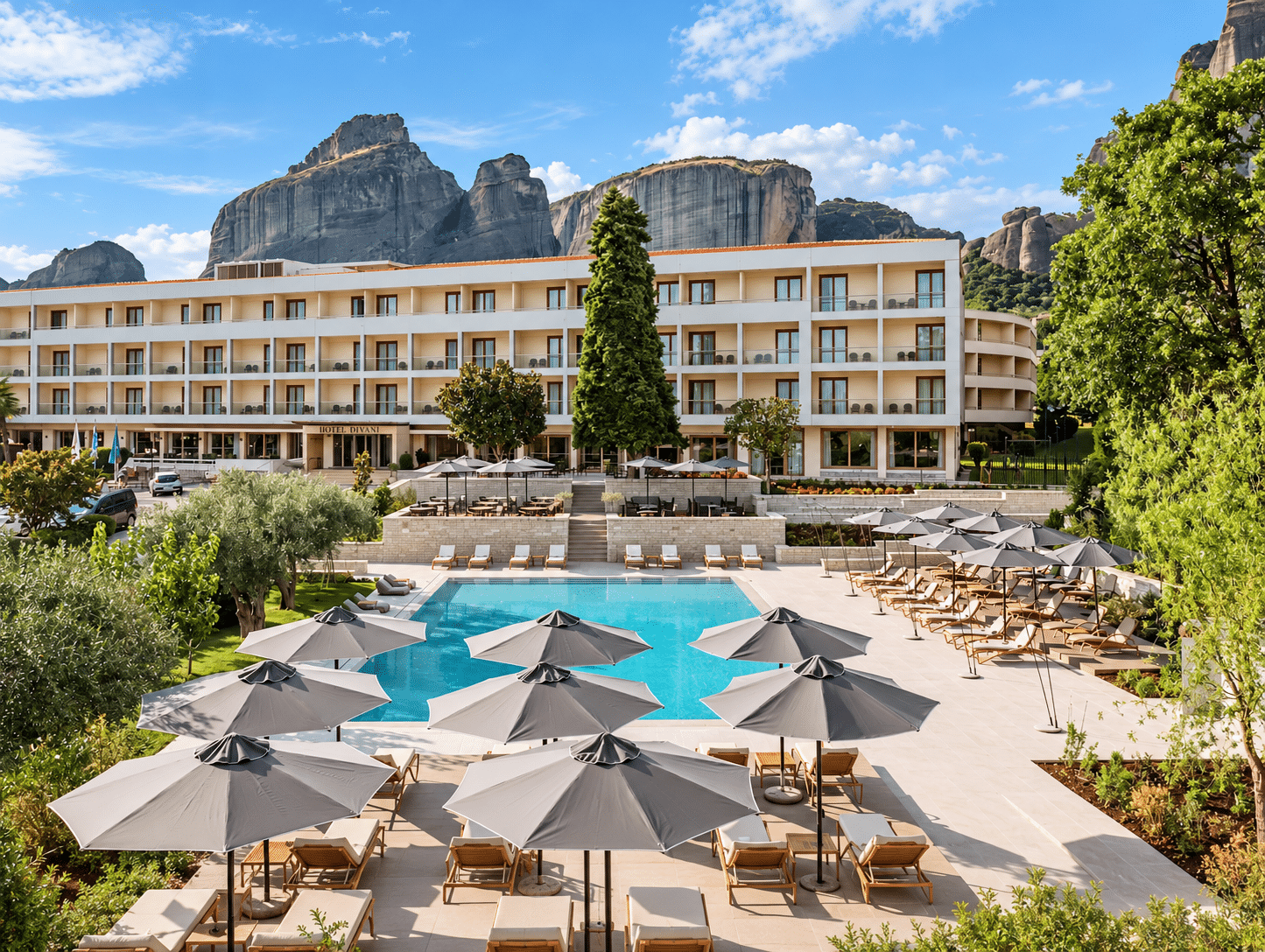 Cream hotel with a blue pool, grey parasols and sun loungers below rocky pillars.