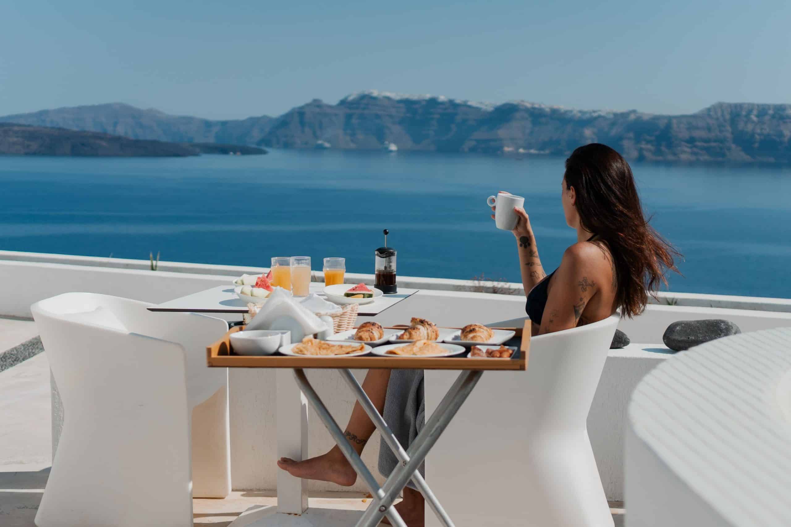 Woman in a bikini at a clifftop breakfast table with coffee and pastries overlooking the caldera sea.